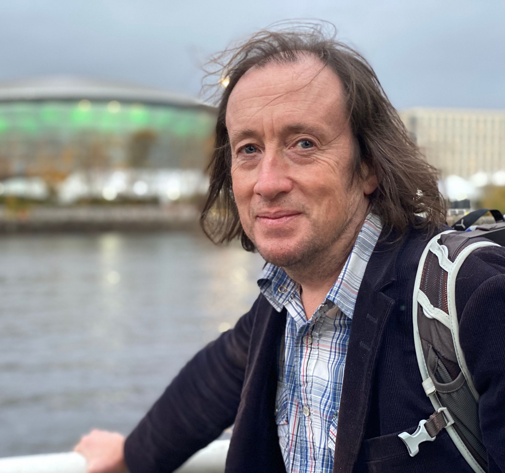 Portrait image of Alasdair standing by the River Clyde, Glasgow. He's holding the safety railing with his right hand. He's wearing a blue-red plaid shirt under his dark blue jacket. In the background the Armadillo is visible..