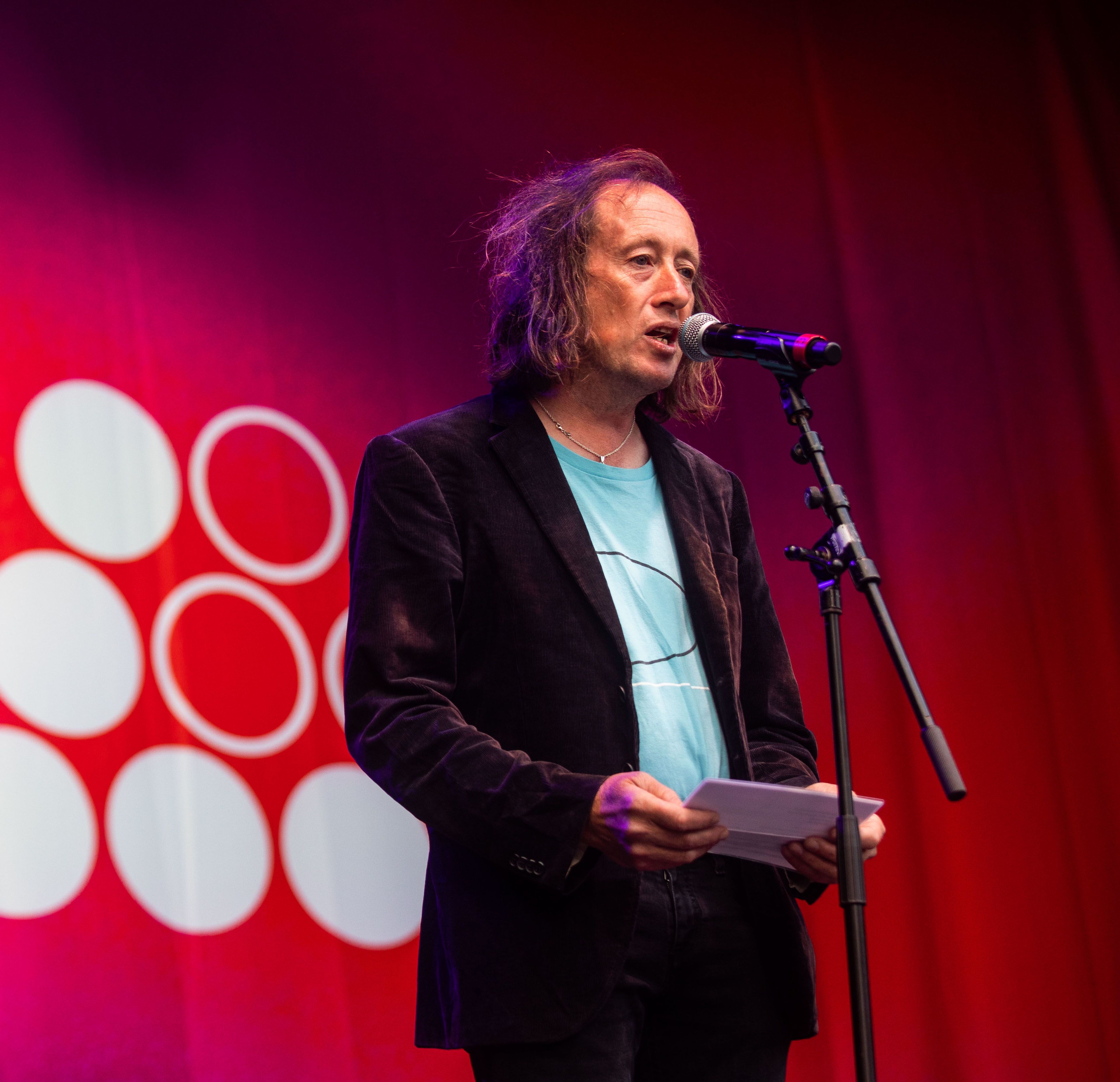 Alasdair standing in front of a microphone, speaking into it as he looks out, behind the camera. In the background, a deep red curtain with the Climate Live logo on it is visible.