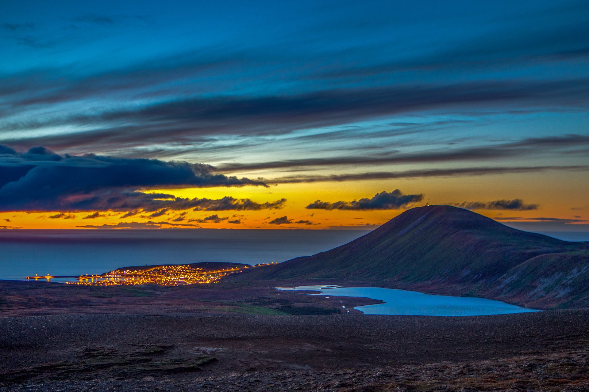 A landscape photograph, out over the shoreline and ocean, with the sunset on the horison. To the right is a large hill and to the left the lights of a small town, just on the shoreline.