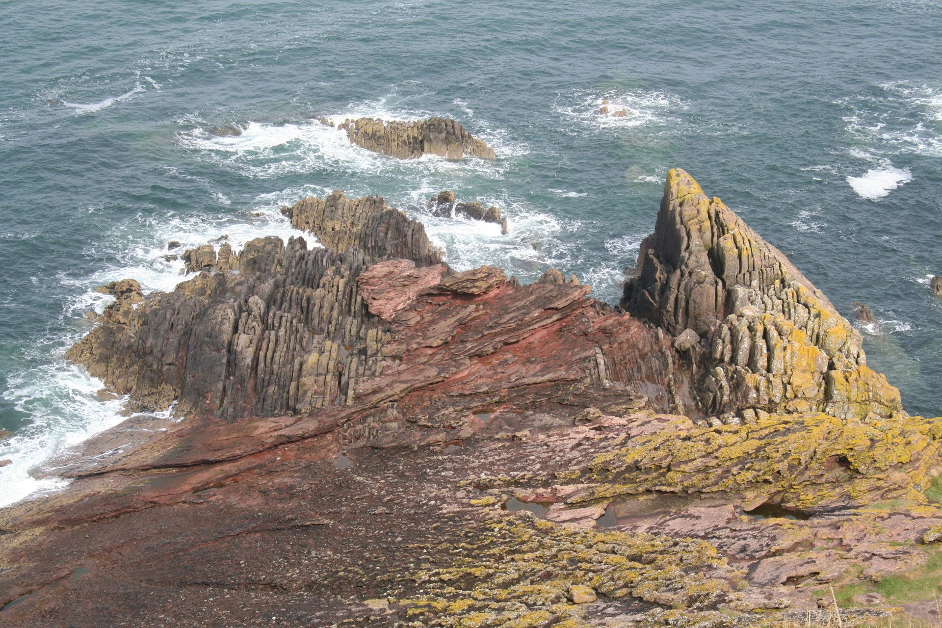 A formation of pointy, green- and red-ish rocks, leading out into the ocean waves.